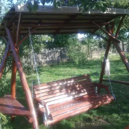 Drewniana huśtawka ogrodowa z zadaszeniem, zawieszona na łańcuchach, na tle zielonego trawnika i drzew owocowych, widoczny fragment drewnianego płotu.