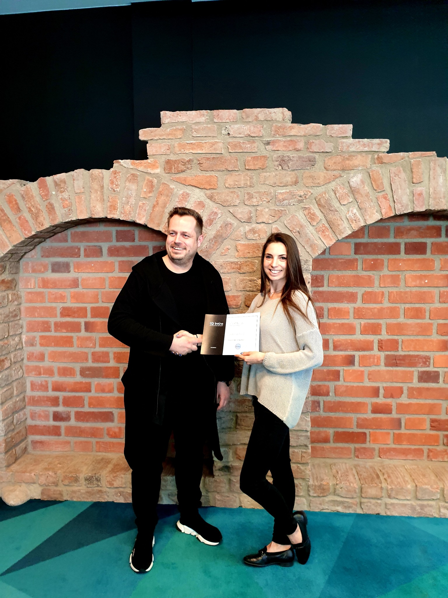 Mężczyzna w czarnym stroju i kobieta w szarym swetrze wymieniają uścisk dłoni, trzymając certyfikat na tle dekoracyjnej ściany z cegły.