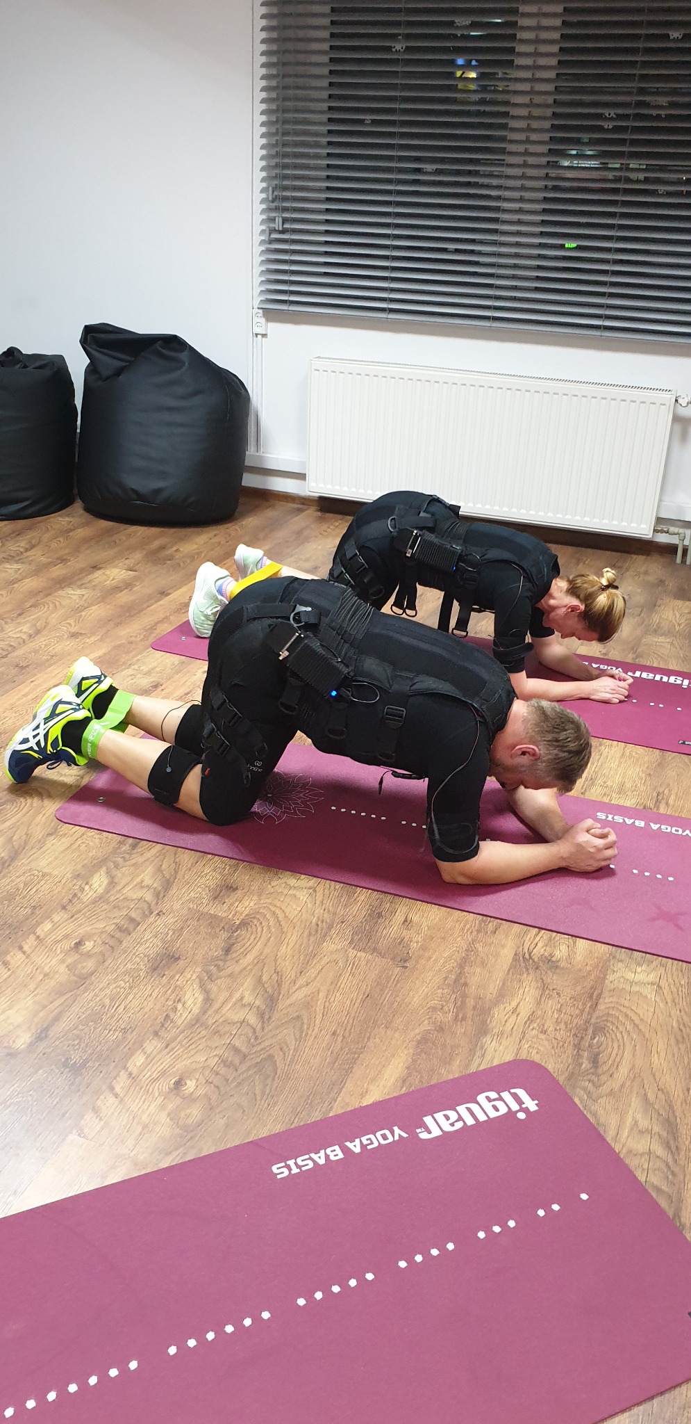 Dwie osoby w kombinezonach EMS ćwiczą plank na matach do jogi w jasnym pomieszczeniu z drewnianą podłogą.