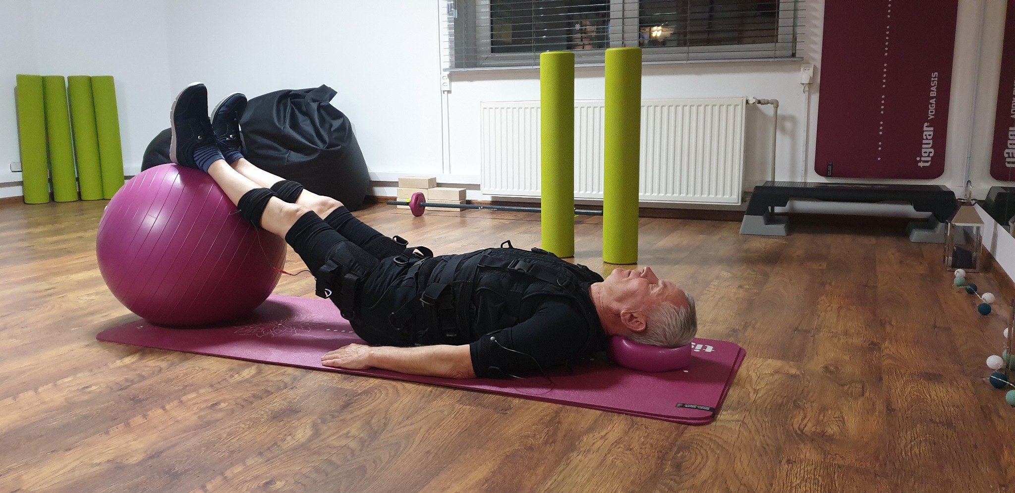 Mężczyzna w kombinezonie EMS leży na macie, z nogami opartymi na fioletowej piłce do ćwiczeń podczas treningu w studiu fitness.