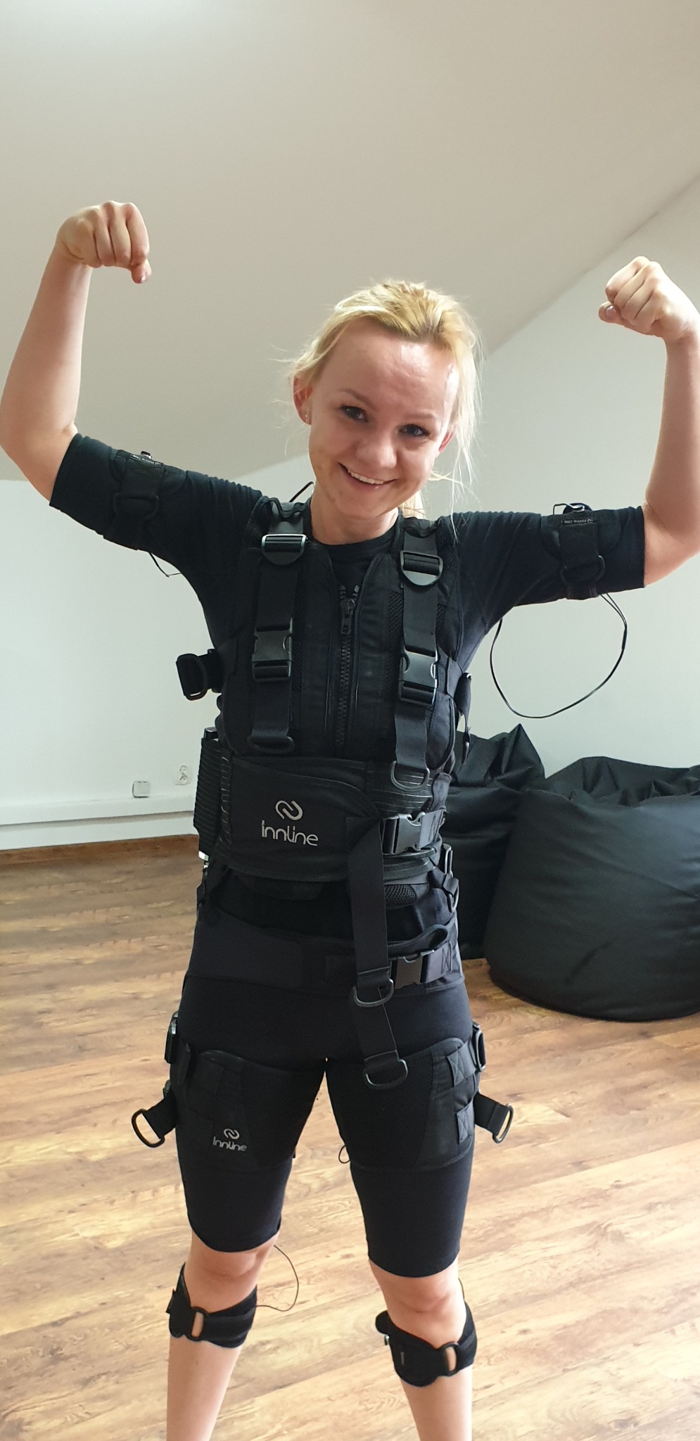 Uśmiechnięta blondynka w czarnym kombinezonie EMS z elektrodami, prezentująca napięte bicepsy na tle jasnej ściany i worków sako.