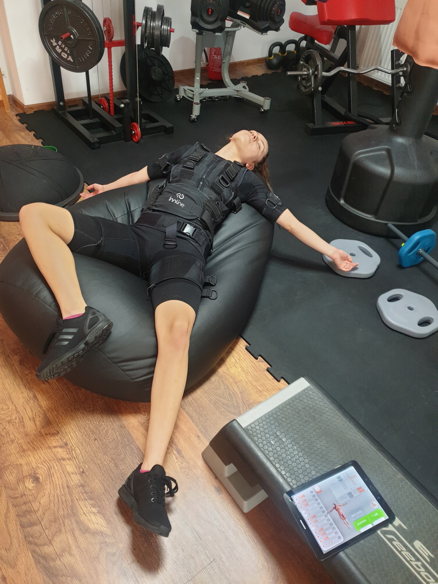 Kobieta w kombinezonie do elektrostymulacji leży na pufie w siłowni, obok tablet z programem treningowym, w tle widoczne ciężary i sprzęt do ćwiczeń.