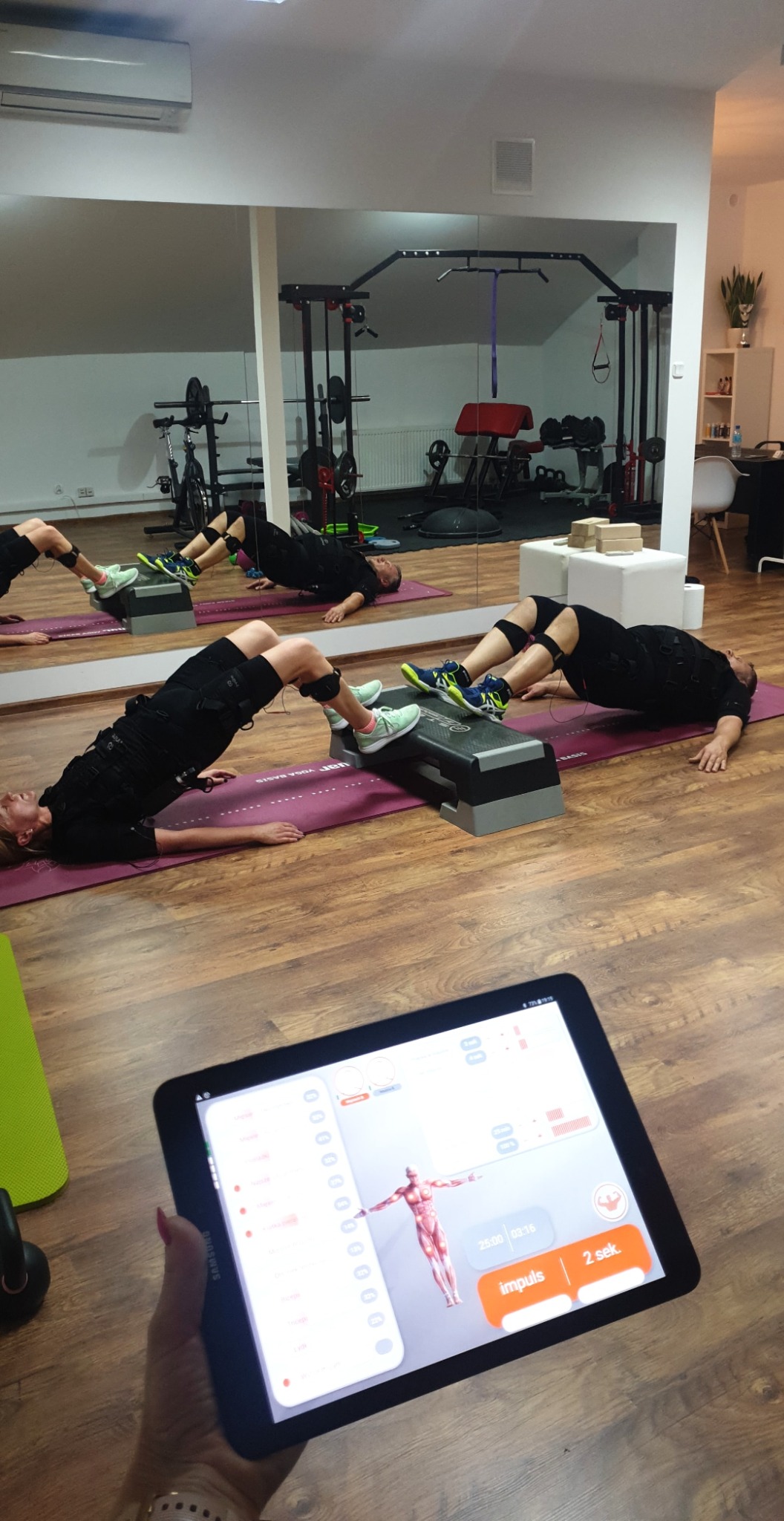 Dwie osoby w czarnych kombinezonach EMS wykonują ćwiczenie unoszenia bioder na stepach w siłowni, tablet w dłoni pokazuje parametry impulsu elektrycznego.