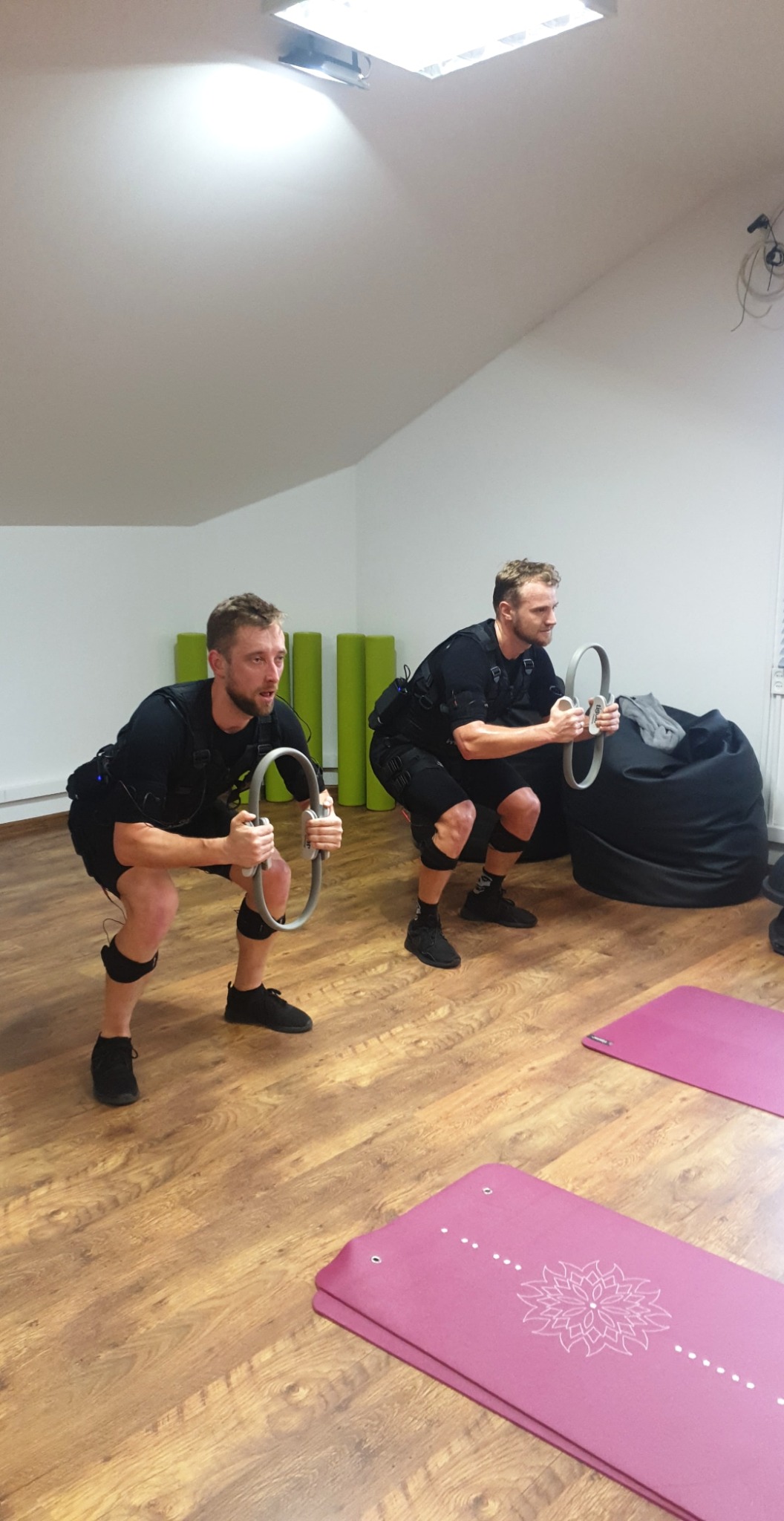 Dwóch mężczyzn w czarnych strojach EMS wykonuje przysiady z pierścieniami, widoczne maty do ćwiczeń i worki sako w tle.