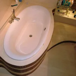 Nowoczesna, owalna wanna z chromowaną armaturą, obudowana płytkami mozaikowymi w odcieniach brązu i beżu, z widoczną półką z kosmetykami w tle.