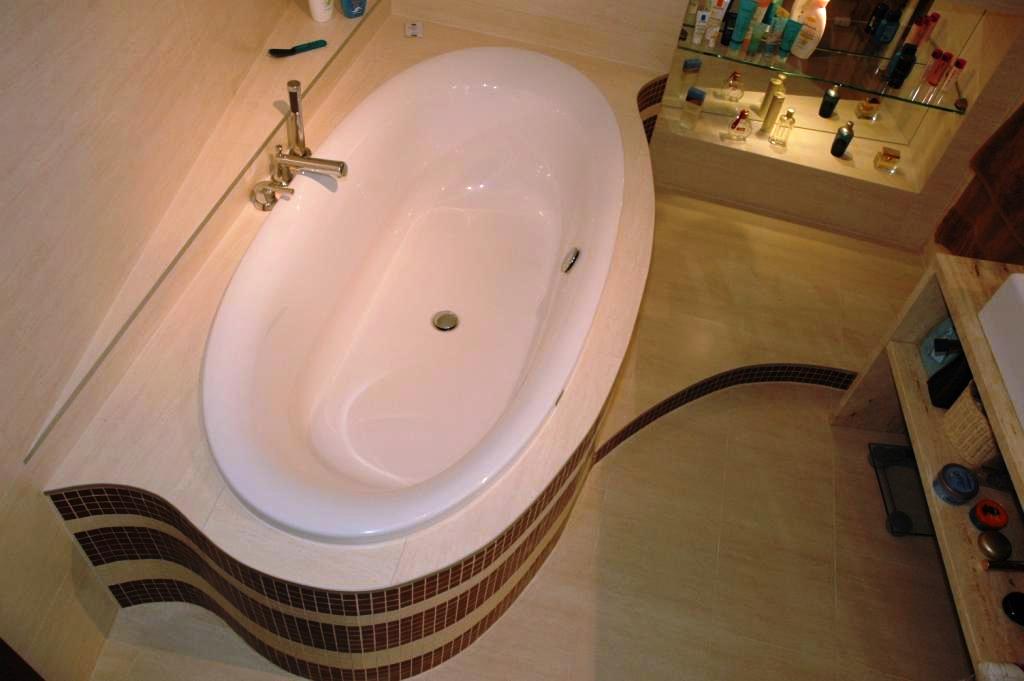 Nowoczesna, owalna wanna z chromowaną armaturą, obudowana płytkami mozaikowymi w odcieniach brązu i beżu, z widoczną półką z kosmetykami w tle.