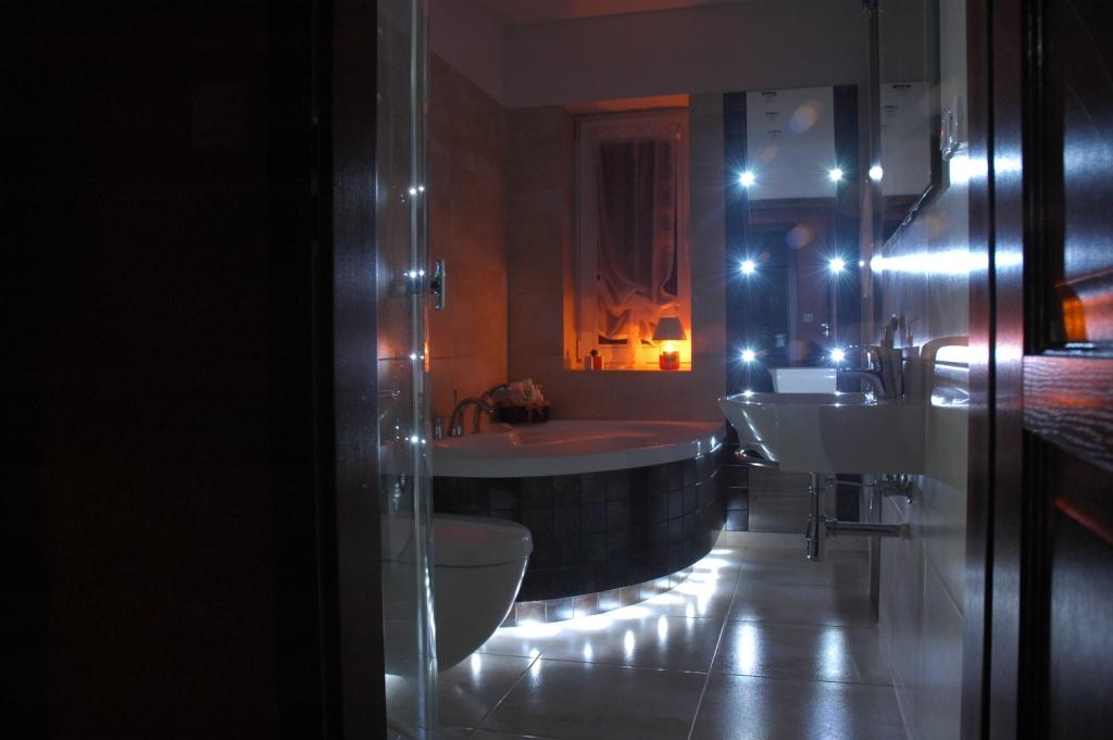 Nowoczesna łazienka z okrągłą wanną obłożoną ciemną mozaiką, podświetleniem LED pod wanną i umywalką na metalowym stelażu, widok zza częściowo otwartych drzwi kabiny prysznicowej.