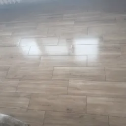 Ułożona podłoga z płytek imitujących drewno, widoczny fragment grzejnika i resztki zaprawy w rogu pomieszczenia.