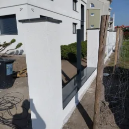 Nowoczesne ogrodzenie z białymi, teksturowanymi słupkami i czarnymi, błyszczącymi elementami wykończeniowymi, w tle biały budynek z szarymi oknami i fragmentem elewacji w odcieniach zieleni.