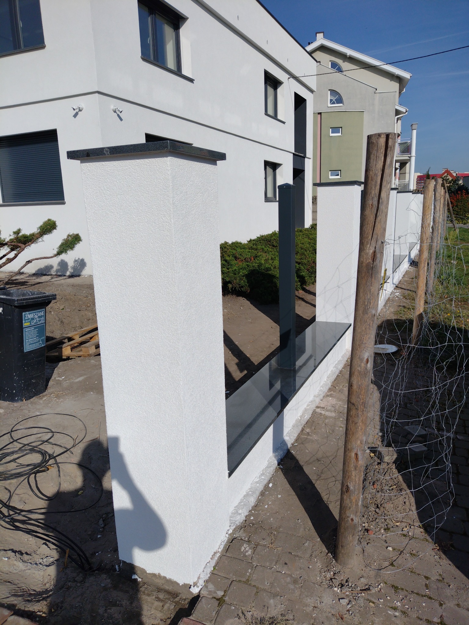 Nowoczesne ogrodzenie z białymi, teksturowanymi słupkami i czarnymi, błyszczącymi elementami wykończeniowymi, w tle biały budynek z szarymi oknami i fragmentem elewacji w odcieniach zieleni.