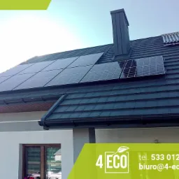 Panele na dachu domu z ciemną dachówką, widoczny komin i kolektory słoneczne. W rogu logo firmy.