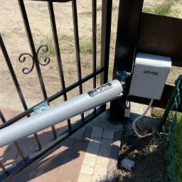 Szary siłownik FAAC zamontowany na czarnej, metalowej bramie posesji, widoczna skrzynka sterownicza i kostka brukowa w tle.