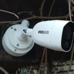 Biała kamera CCTV zamontowana na ścianie, otoczona suchymi gałęziami i z logotypem firmy 'bixit' w prawym dolnym rogu.