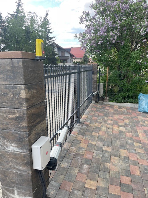 Automatyczna brama wjazdowa z czarnymi, kutymi przęsłami, zamontowana na kamiennych słupkach. Widoczny napęd bramy i lampa ostrzegawcza. Kostka brukowa na podjeździe.