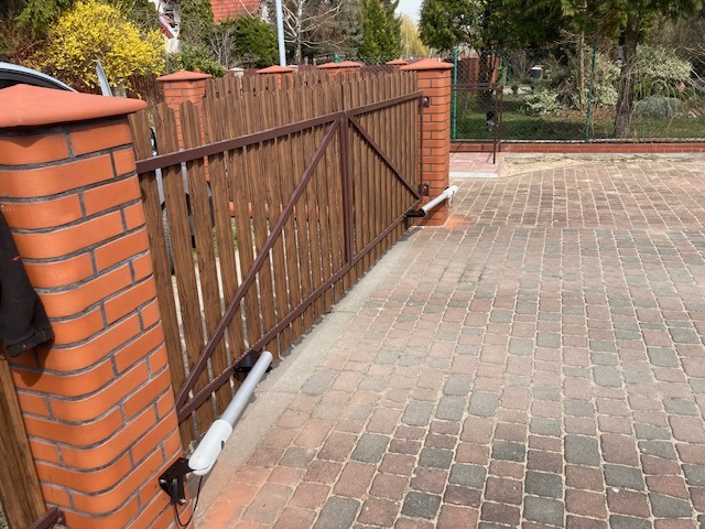 Automatyczna brama posesji z brązowego drewna i ceglanymi słupkami, widoczny napęd i kostka brukowa na podjeździe. Słoneczny dzień.