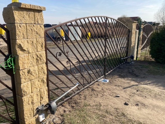 Kuta brama wjazdowa z automatyką, osadzona na kamiennych słupkach. Widoczny mechanizm otwierający bramę. Tło: zabudowa jednorodzinna i błękitne niebo.