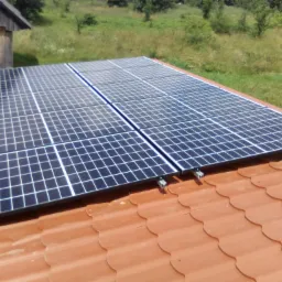 Panele słoneczne zamontowane na dachu z czerwonej dachówki, widok z góry, w tle zieleń drzew i krzewów.