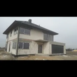 Dwukondygnacyjny dom w stanie surowym otwartym, z widocznymi oknami, czarną bramą garażową i fragmentami ocieplenia fundamentów, na tle pochmurnego nieba i niezagospodarowanego terenu.