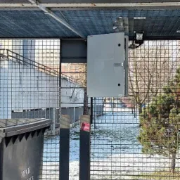 Szara szafka sterownicza zamontowana pod zadaszeniem z kraty, prawdopodobnie do autonomicznego oświetlenia wiat śmietnikowych w Katowicach, widoczny kosz na śmieci z napisem MPGk Katowice.