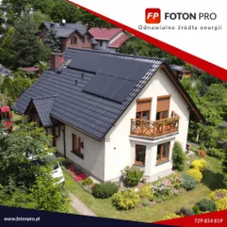 Dom jednorodzinny z ciemną dachówką i panelami słonecznymi na dachu, widziany z góry, z zadbanym ogrodem i drewnianym balkonem.