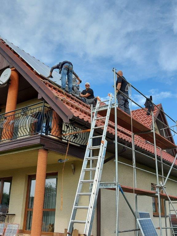 Montaż paneli słonecznych na dachu domu jednorodzinnego w Rusocicach: ekipa instalatorów na rusztowaniu i drabinie, częściowo zamontowane panele na czerwonej dachówce, błękitne niebo w tle.