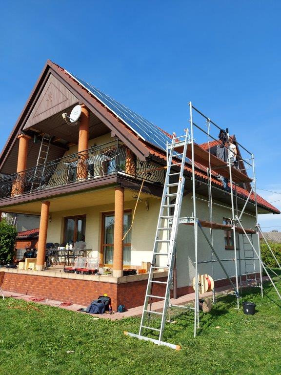 Montaż paneli na dachu dwupiętrowego domu w Rusocicach, widoczne rusztowanie i pracownicy w trakcie instalacji, błękitne niebo.