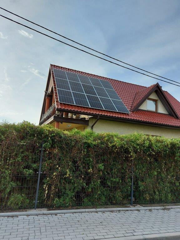 Panele słoneczne na dachu dwupiętrowego domu z czerwonej dachówki, widoczne zza żywopłotu.