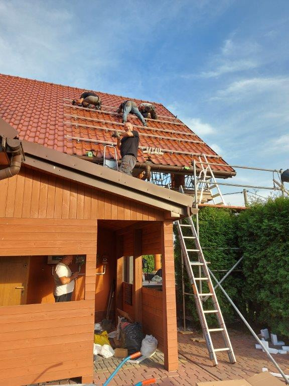 Montaż konstrukcji wsporczej na dachu z czerwonej dachówki, przygotowanie do instalacji paneli słonecznych, widoczni monterzy na dachu i drabina oparta o budynek.