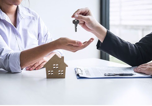 Przekazanie kluczy do domu: dłoń w ciemnym garniturze wręcza klucz osobie w białej koszuli, na stole drewniana figurka domu i umowa na podkładce z klipsem.