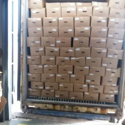 Wnętrze kontenera wypełnione kartonami z otworami wentylacyjnymi, ułożonymi warstwami od podłogi do sufitu; widoczne elementy konstrukcyjne kontenera.