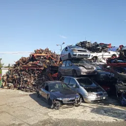 Stos zdemontowanych samochodów i części samochodowych na placu składowym, z widocznym czerwonym pojazdem w tle i błękitnym niebem.