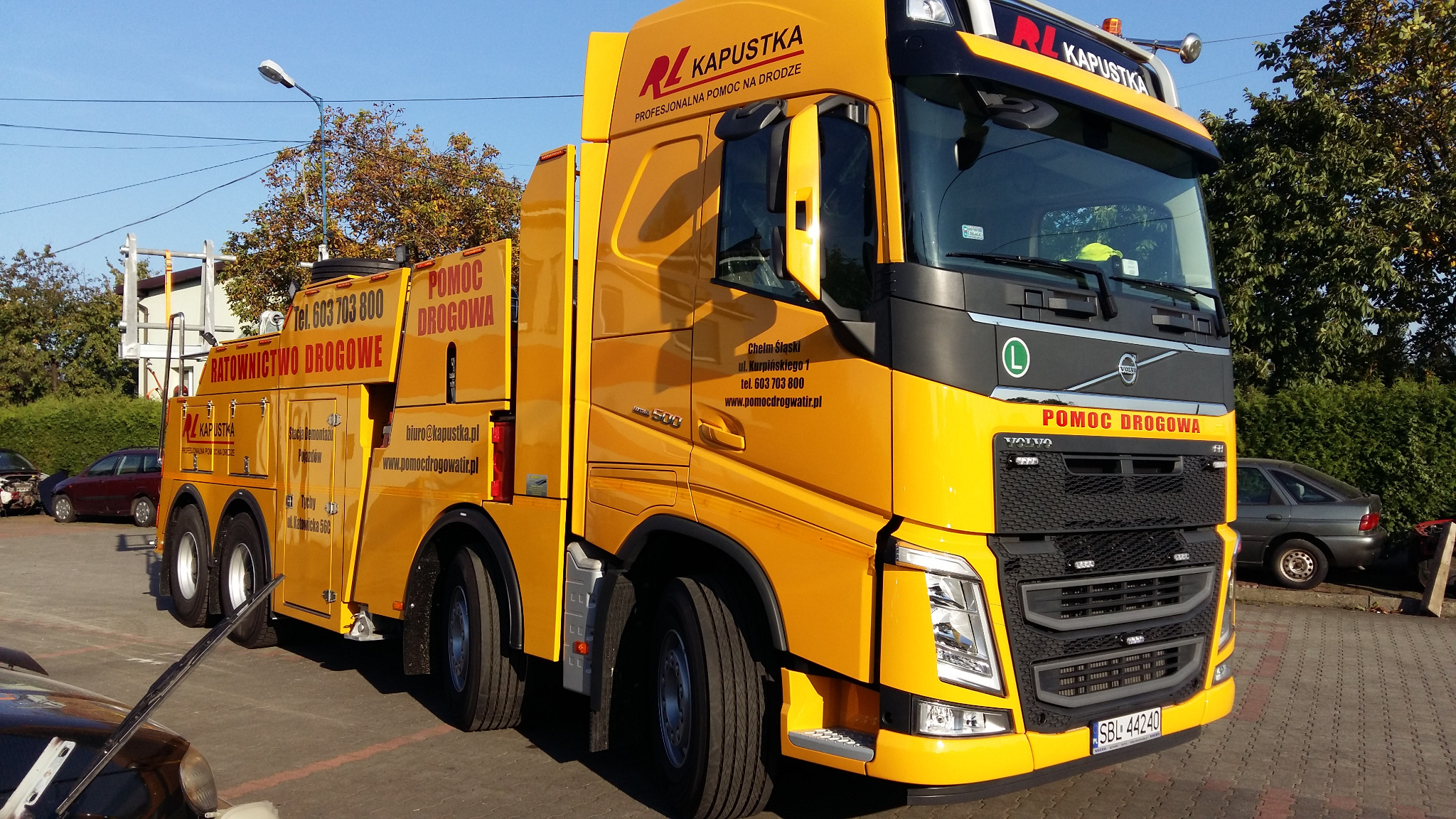 Żółty samochód pomocy drogowej marki Volvo z napisem 'POMOC DROGOWA' i danymi kontaktowymi firmy 'Kapustka' na parkingu.