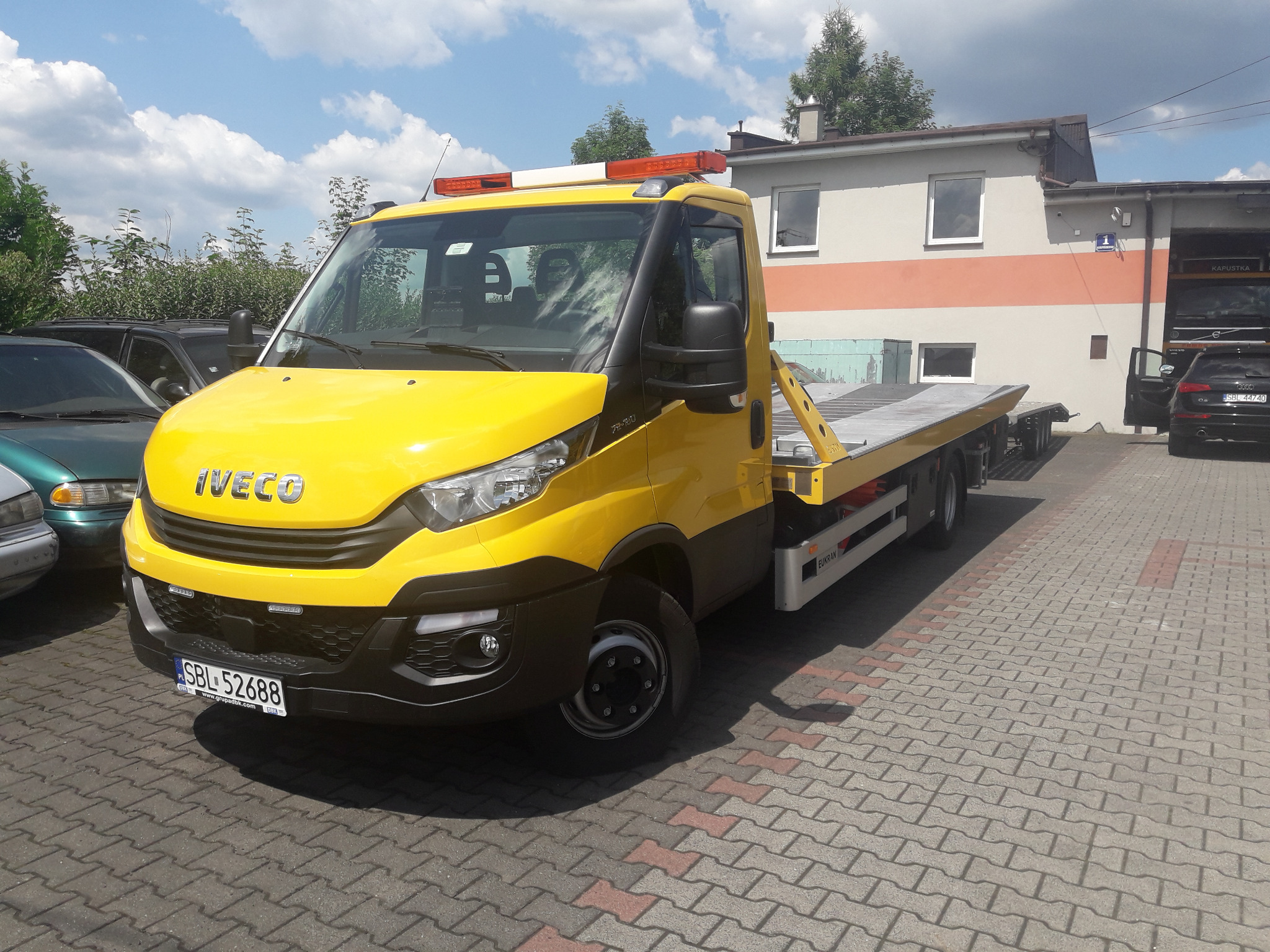 Żółta laweta Iveco z pomarańczowym światłem ostrzegawczym, zaparkowana na kostce brukowej przed budynkiem warsztatu.