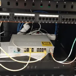 Panel krosowniczy Cat.6 z podłączonymi kablami światłowodowymi i patchcordami, switch Cisco Catalyst 2960-L Series, listwa zasilająca i router w szafie rack