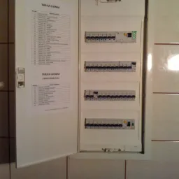 Otwarta skrzynka rozdzielcza z listwą zaciskową i naklejoną listą obwodów na tle kafelków.