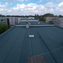 Zielony dach z papy termozgrzewalnej na bloku, widok z góry na tle zabudowy miejskiej i błękitnego nieba z chmurami.