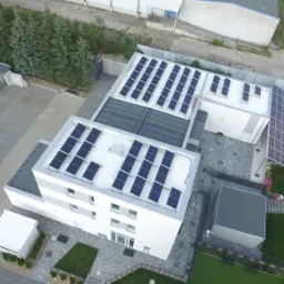 Widok z drona na budynek z panelami słonecznymi na dachu i w formie zadaszenia nad wejściem, obok parking z zaparkowanym czarnym samochodem.