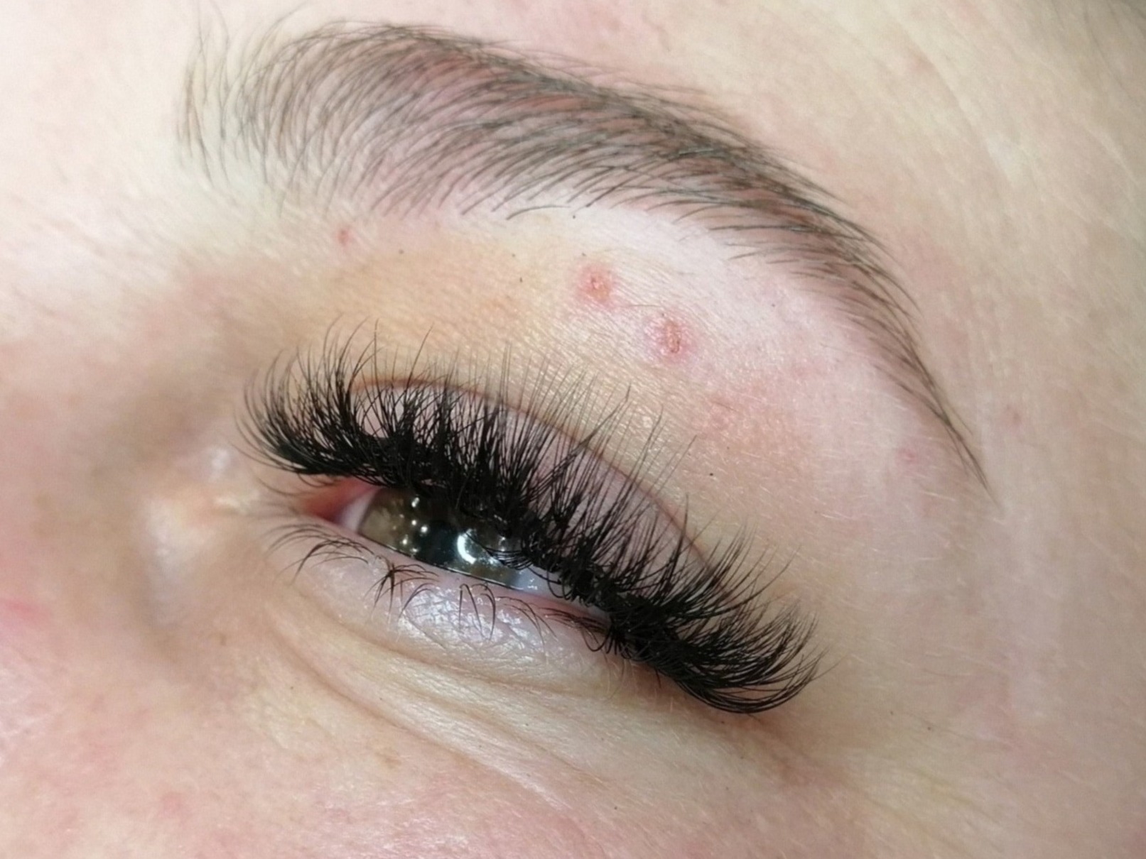 Zbliżenie na oko z przedłużonymi, gęstymi, czarnymi rzęsami, widoczne drobne niedoskonałości skóry nad powieką.