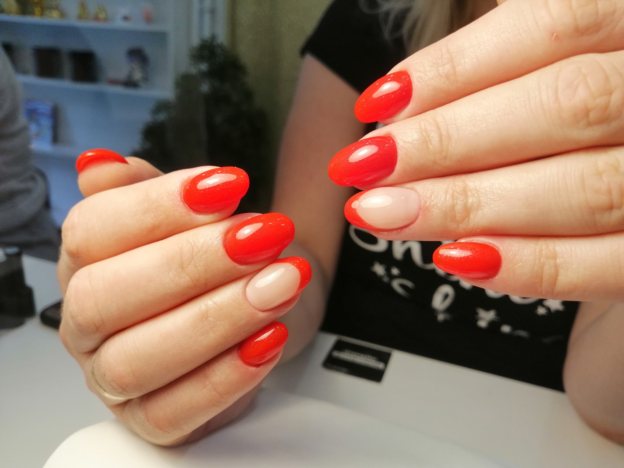 Dłonie z czerwonym, błyszczącym manicure hybrydowym, dwa paznokcie z french manicure na beżowym tle, owalny kształt paznokci.
