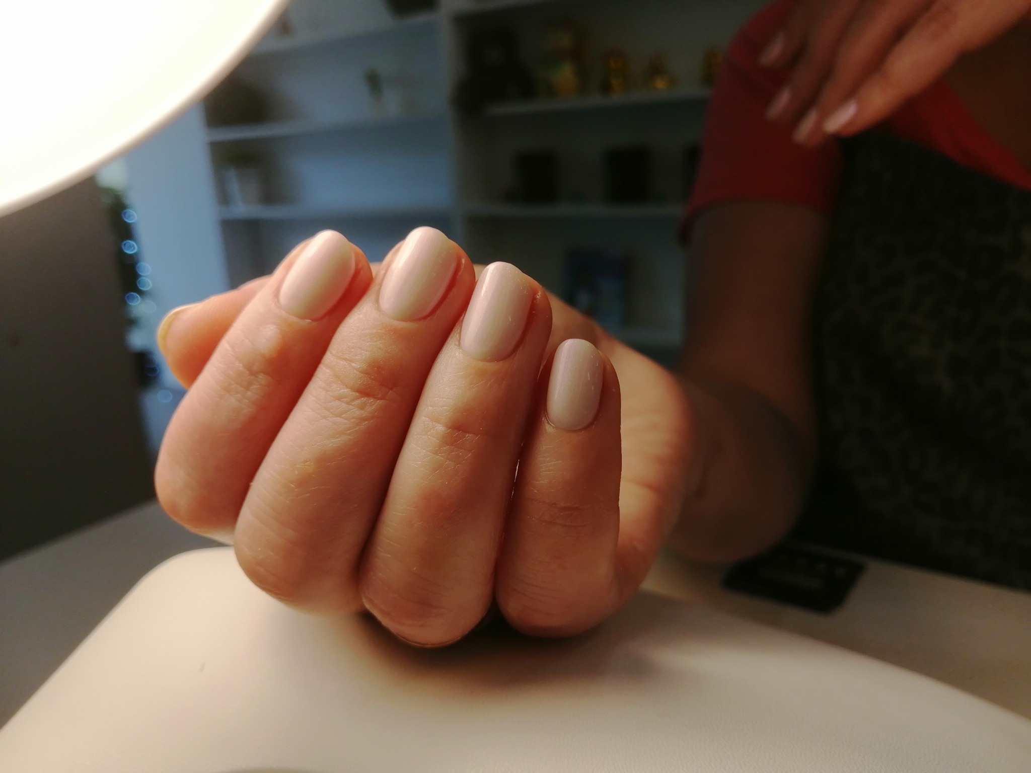 Dłoń z paznokciami pomalowanymi na jasny, kremowy kolor, widoczne delikatne skórki, w tle regał z kosmetykami i lampa do utwardzania lakieru hybrydowego.