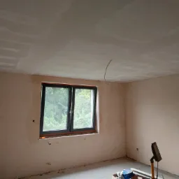 Pokój w trakcie prac wykończeniowych: ściany i sufit pokryte warstwą szpachli, widoczne ślady po łatach na suficie, okno z widokiem na zieleń, na podłodze narzędzia i materiały.