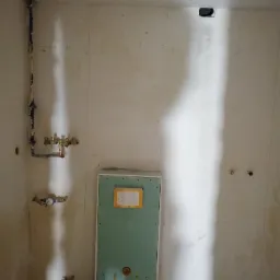 Surowa łazienka w trakcie remontu: widoczne rury instalacji wodnej, zabudowa stelaża podtynkowego WC z płyt karton-gips oraz odpływ liniowy w podłodze. Ściany przygotowane do dalszych prac...