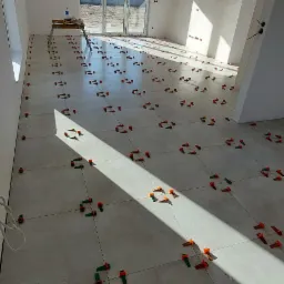 Jasne płytki podłogowe w trakcie układania, z widocznymi klipsami poziomującymi i krzyżykami dystansowymi, w nowym, pustym pomieszczeniu z dużym oknem balkonowym.