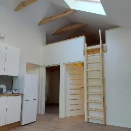 Jasne wnętrze mieszkania po remoncie z białą zabudową kuchenną, lodówką, drewnianą drabiną prowadzącą na antresolę i oknem dachowym.