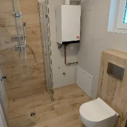 Nowoczesna łazienka z kabiną prysznicową typu walk-in, wiszącą toaletą i piecem gazowym na ścianie, wykończona płytkami imitującymi drewno.