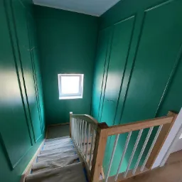 Klatka schodowa w trakcie malowania na intensywny odcień zieleni, zabezpieczone schody, drewniana balustrada z białymi szczeblami, dekoracyjne listwy ścienne.