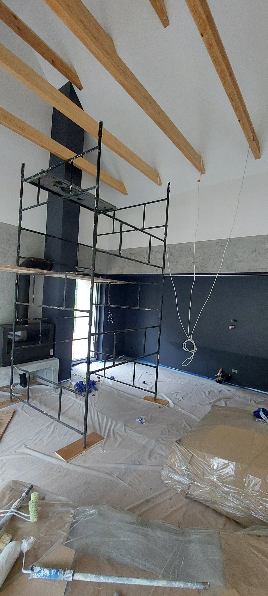 Wnętrze w trakcie malowania na ciemny kolor, widoczne rusztowanie, drewniane belki stropowe i zabezpieczona podłoga z narzędziami malarskimi.