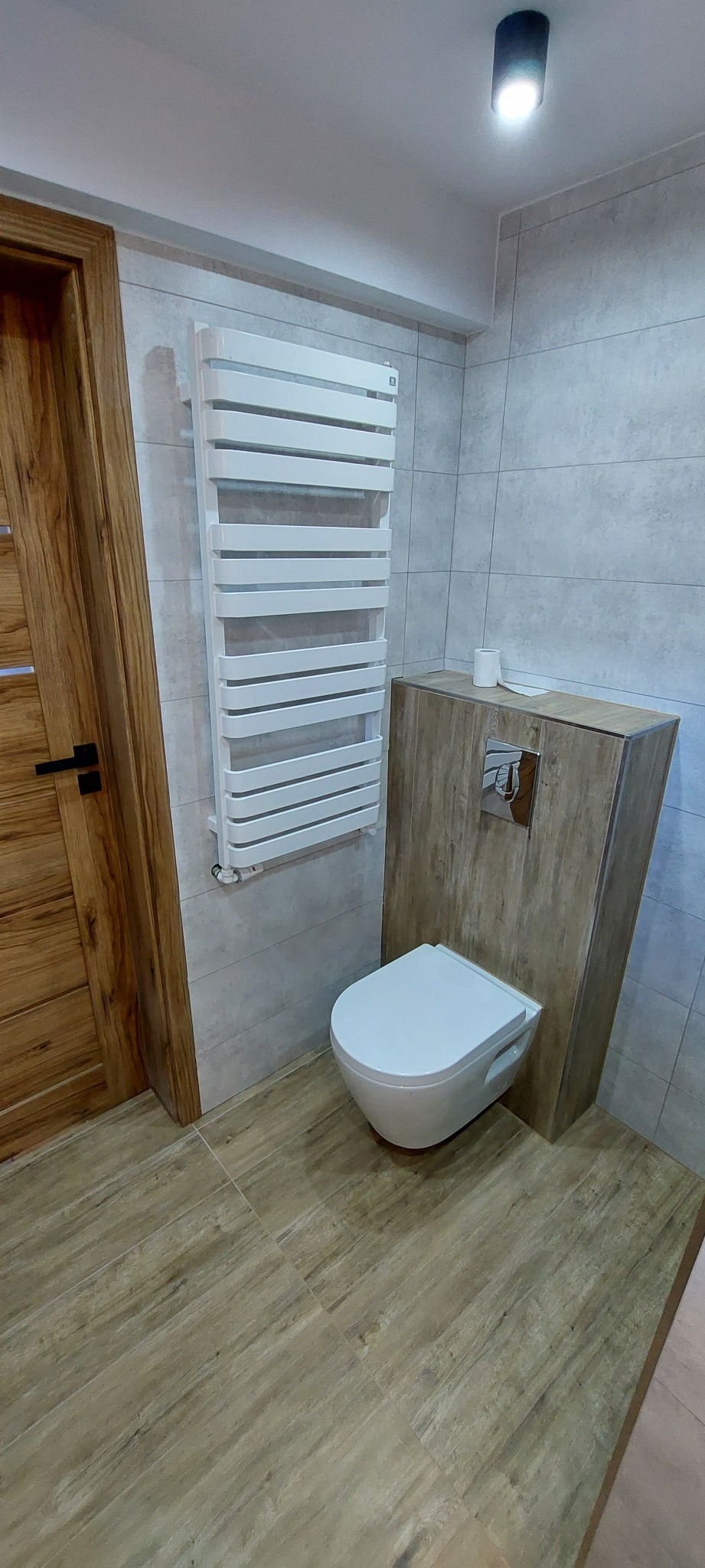 Nowoczesna łazienka z podwieszaną miską WC obudowaną płytami imitującymi drewno, grzejnikiem drabinkowym i płytkami w odcieniach szarości.