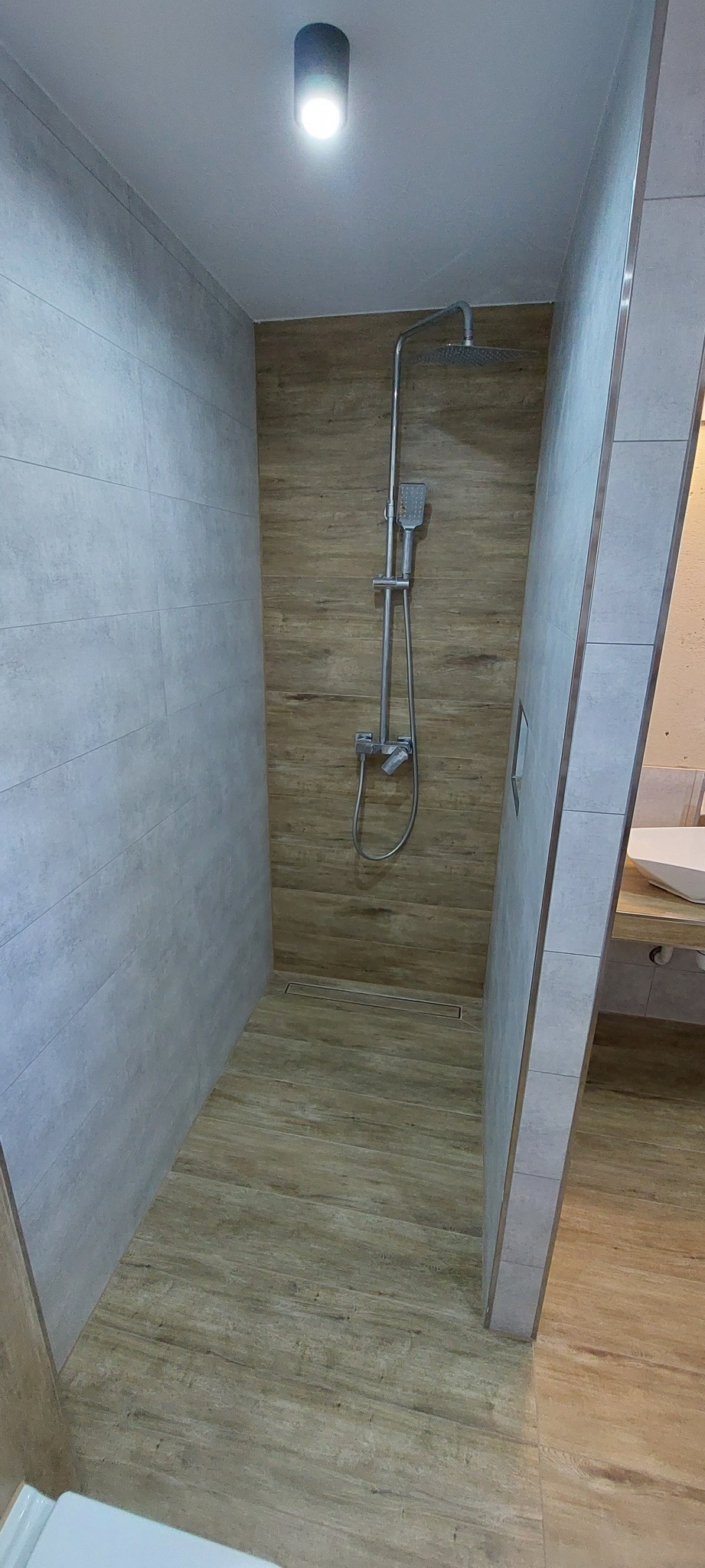 Nowoczesny prysznic typu walk-in z odpływem liniowym, wykończony płytkami imitującymi drewno i szare płytki ścienne.