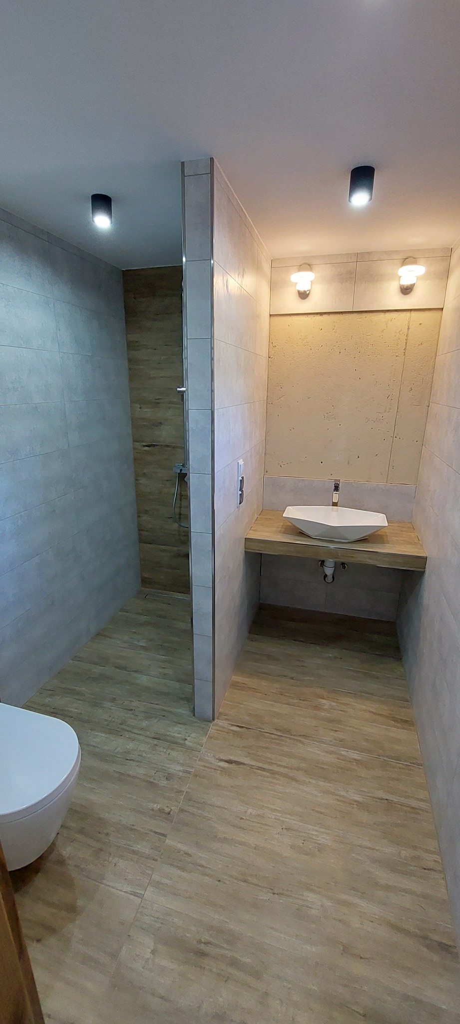 Nowoczesna łazienka z kabiną prysznicową typu walk-in, umywalką nablatową o geometrycznym kształcie i drewnopodobnymi płytkami na podłodze i ścianach.